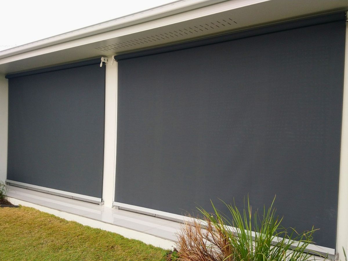 Close-up of a premium cable-guided outdoor blind wire system anchored with a stainless steel L-bracket and hex screws to a modern light grey rendered exterior wall underneath a window.