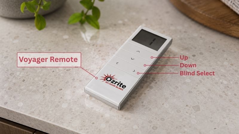 Ozrite Automation Voyager multi-channel outdoor blind remote control resting on a modern stone kitchen benchtop, featuring labeled diagram lines pointing to the LCD screen, up button, down button, and blind select control.